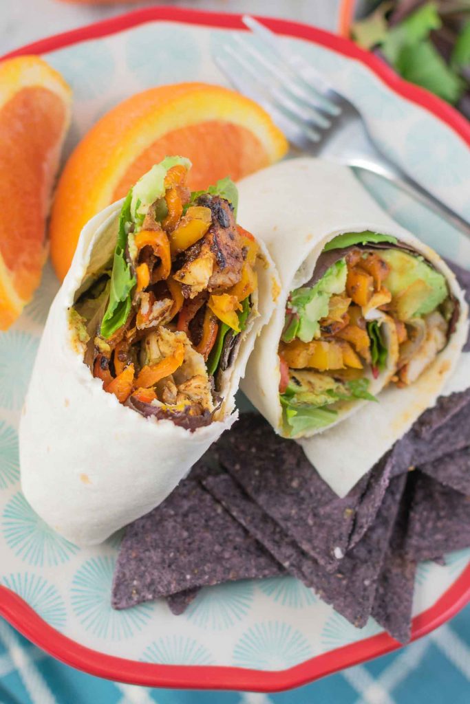 grilled chicken strips, avocado, bacon and veggies in a wrap on a plate with blue chips