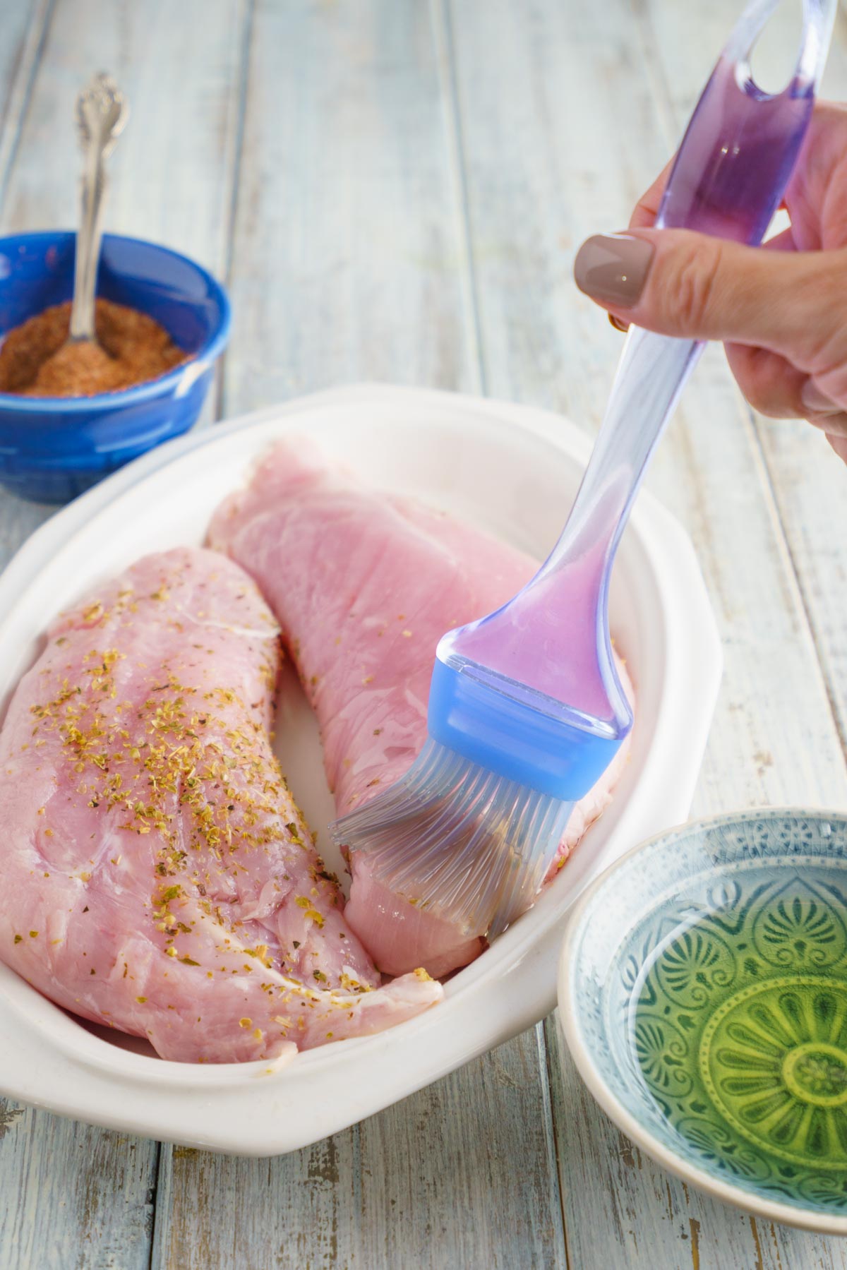 a blue silicone brush bastes poultry with a small bowl of olive oil and a rub next to it.