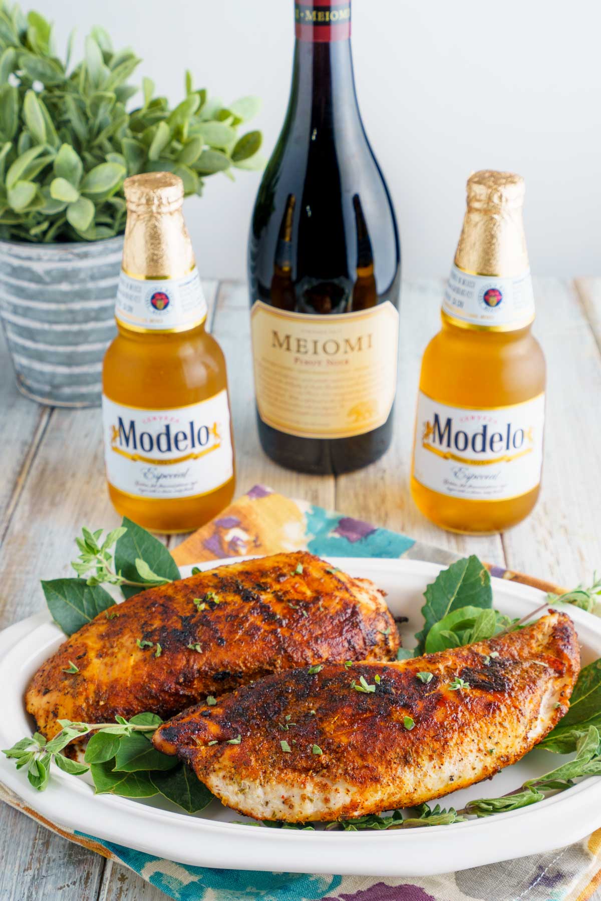 a bottle of red wine and 2 Modelo beers next to a plant and a platter of cooked blackened turkey fillets.