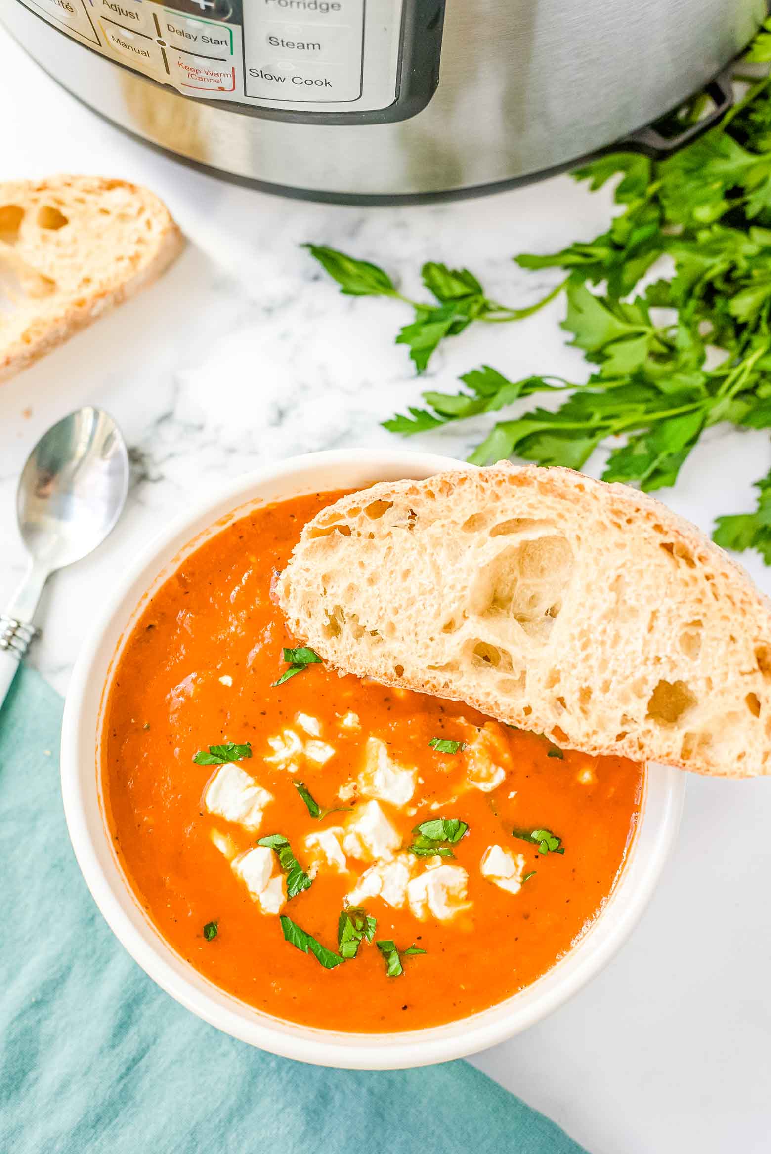 An Instant Pot on a marble countertop with a white spoon, turquoise napkin, fresh parsley and a bowl of pepper and tomato soup with goat cheese