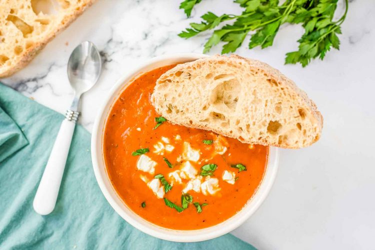 marble countertop with a white spoon, turquoise napkin, fresh parsley and a bowl of pepper and tomato soup with goat cheese