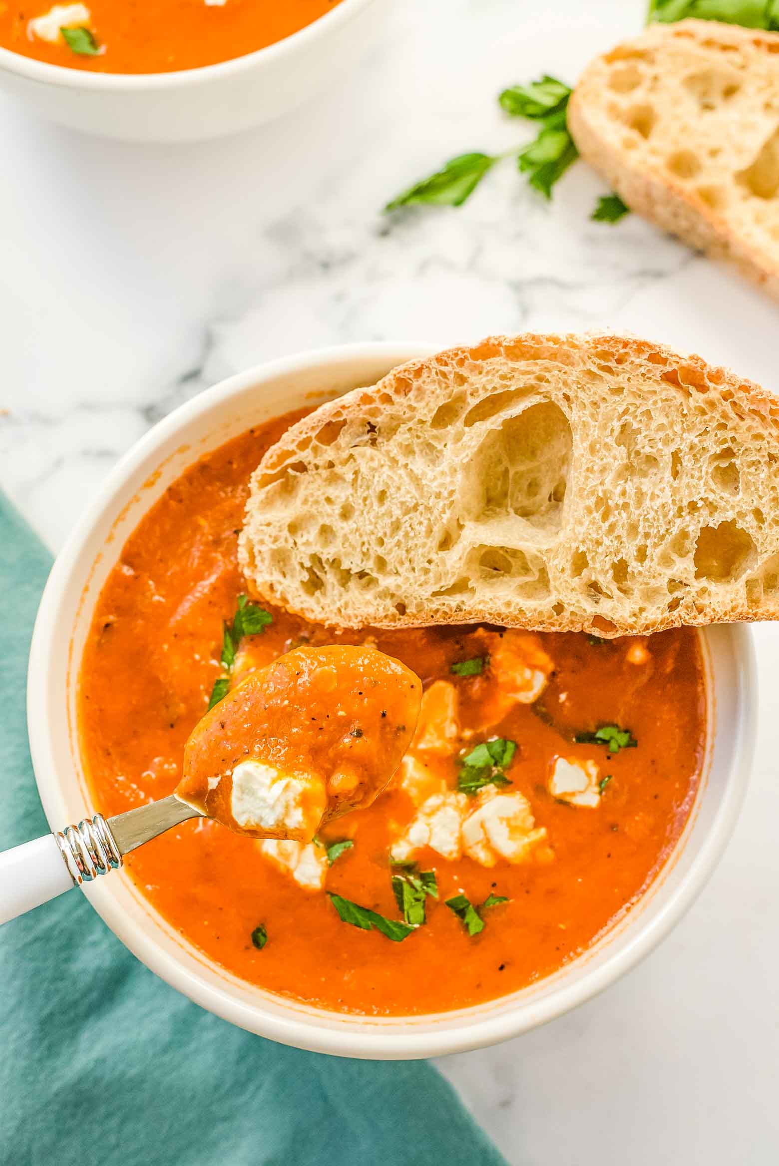 A spoon of soup with cheese and herbs above a full white bowl with a side of french bread