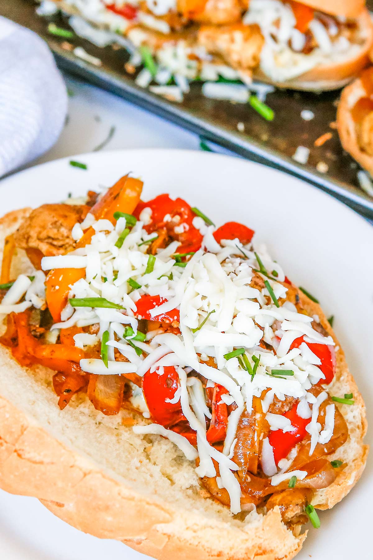 a hoagie bun full of cooked chicken breast pieces, peppers and onions, white shredded cheese and green chives next to a baking pan with several others.