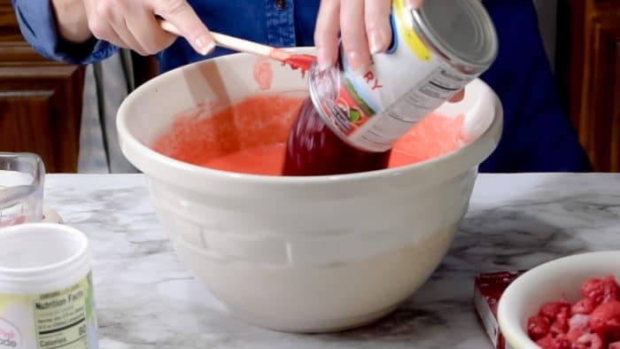 Adding canned cranberry sauce to a white mixing bowl with a red unset jello mixture.