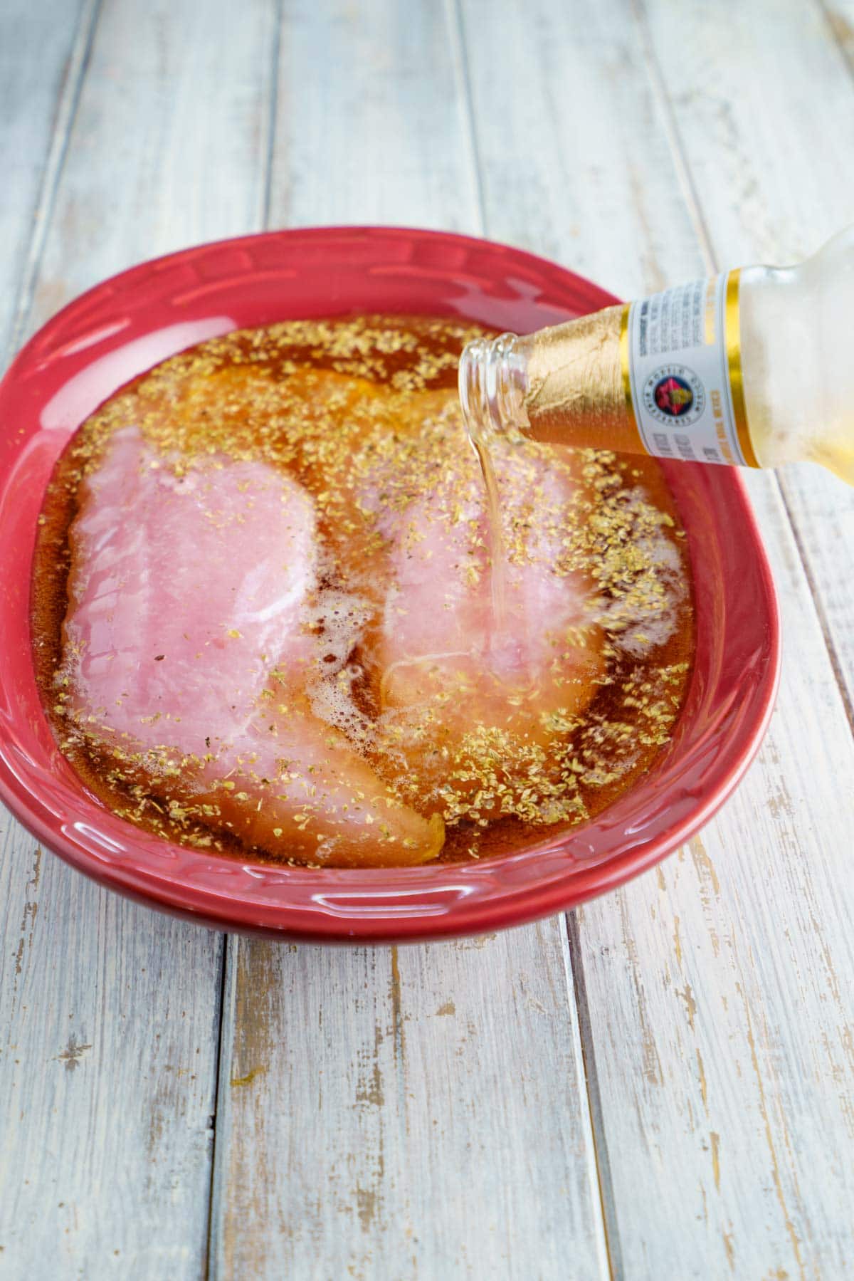 A glass bottle of beer pouring over 2 turkey tenderloins in an oval maroon bowl with spices floating on top.