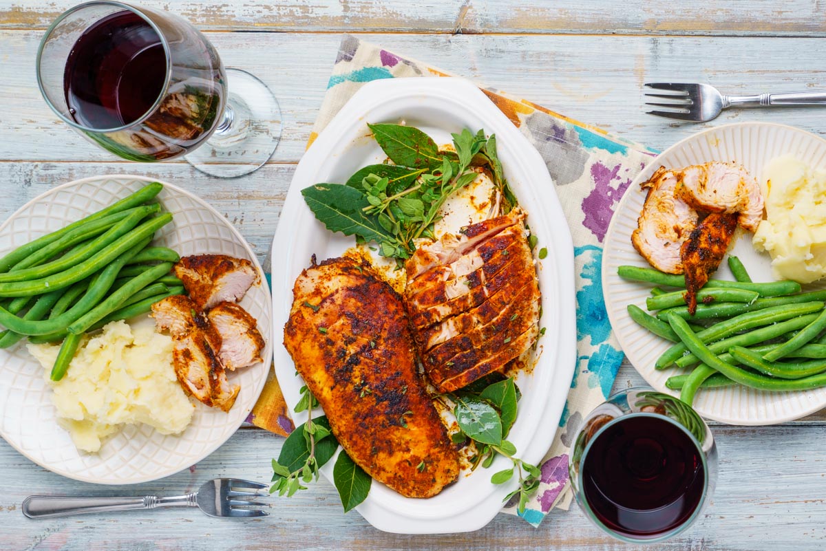 A dinner table set with a platter of grilled blackened turkey tenderloins on a bed of fresh herbs with two dinner plates holding green beans, slices of turkey and mashed potatoes. There are 2 glasses of red wine.