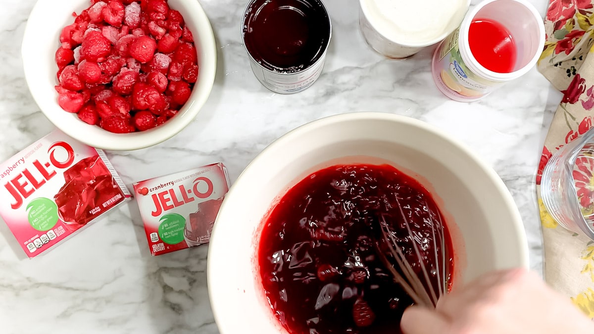 Stirring red jello in a white bowl with a whisk.