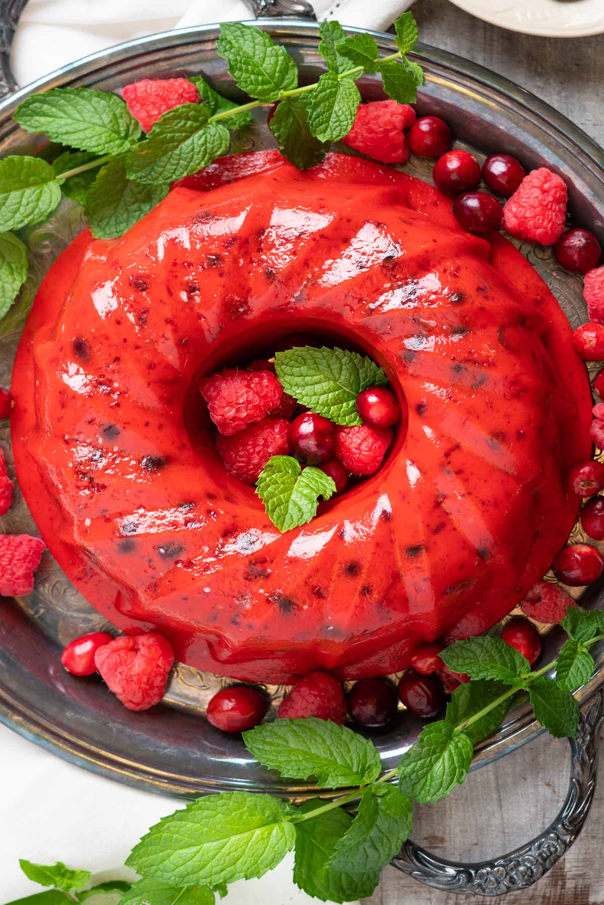 A red jello salad ring with fruit embedded on a tray with fresh fruits and mint.