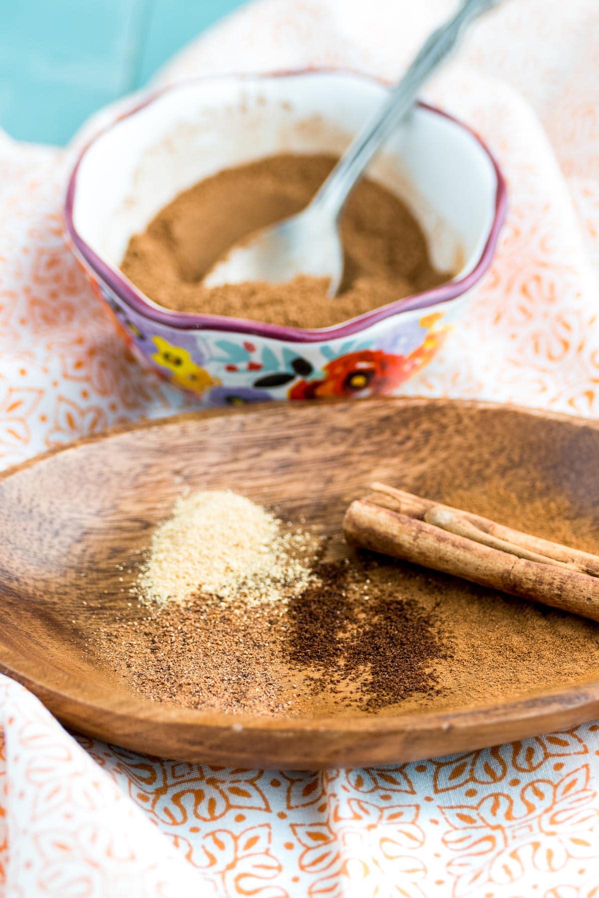 a small bowl of spice mix with a spoon next to a wooden platter that has a cinnamon stick, nutmeg, cloves and ginger. 