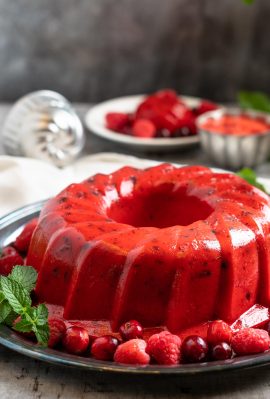 a shiny red jello salad ring sits on a silver platter surrounded by berries and fresh green mint.