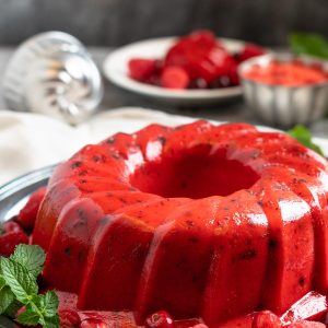 a shiny red jello salad ring sits on a silver platter surrounded by berries and fresh green mint.