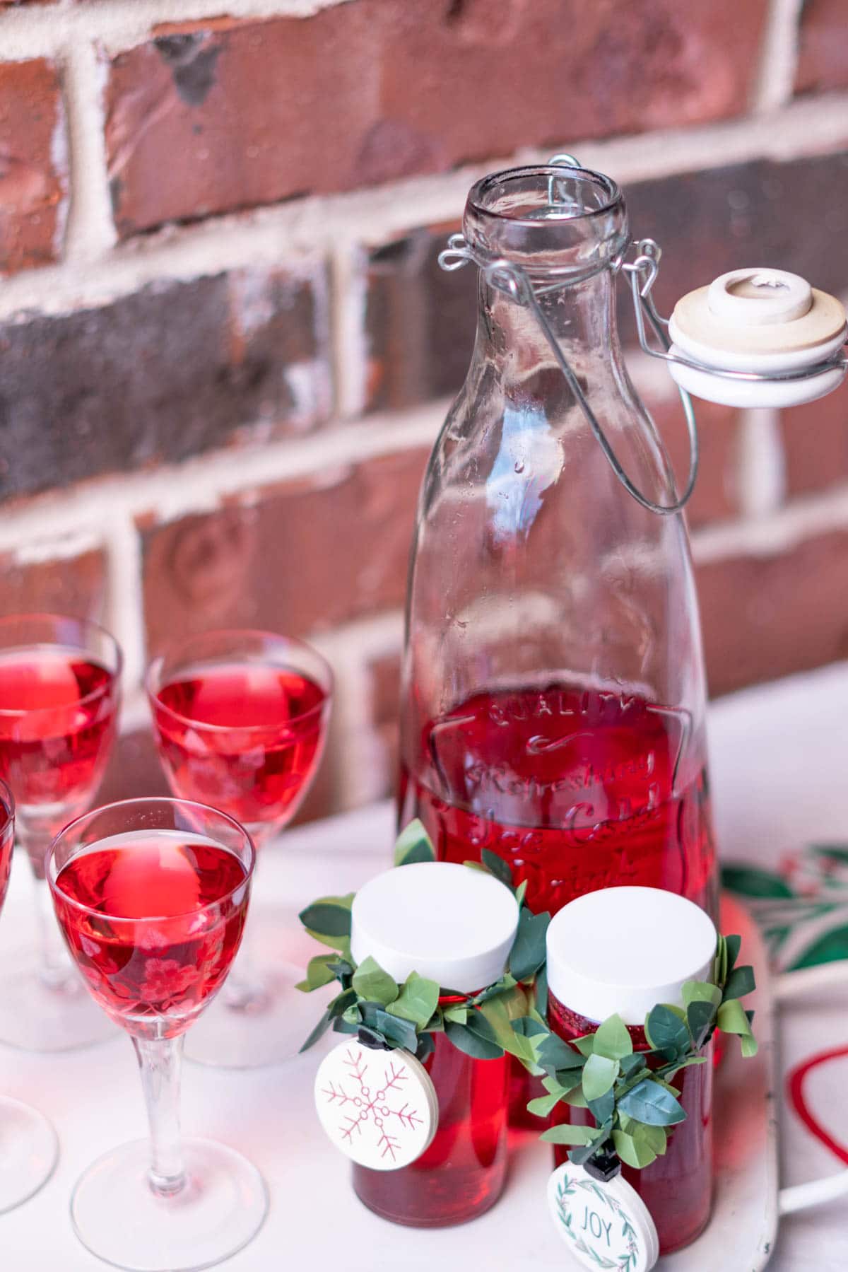 An open glass jar full of red cranberry vodka next to full vintage etched stemmed shot glasses and two smaller bottles meant for gift giving.