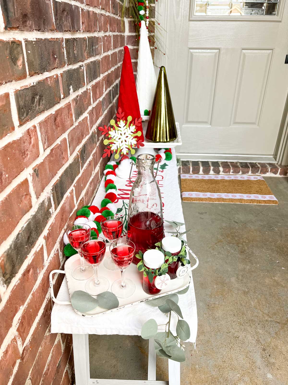 a white bench along a wall decked out for the holidays with a tray of red beverages, colored knit and glass Christmas trees on a cake stand and strings of pom poms.