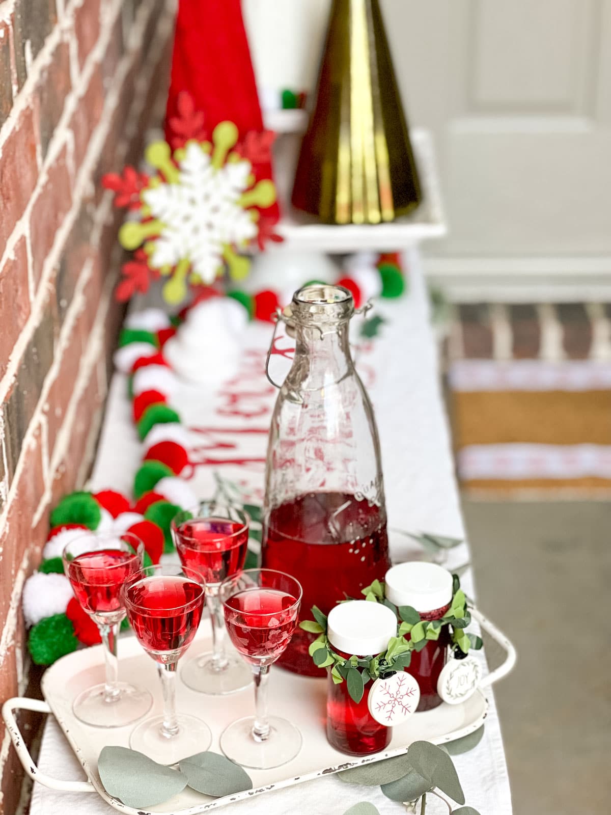 A white metal tray with handles holding 4 cordial glasses and a glass pitcher with homemade cranberry vodka. There are two small glass jars with holiday trim around the neck with small tags as a gift idea. 