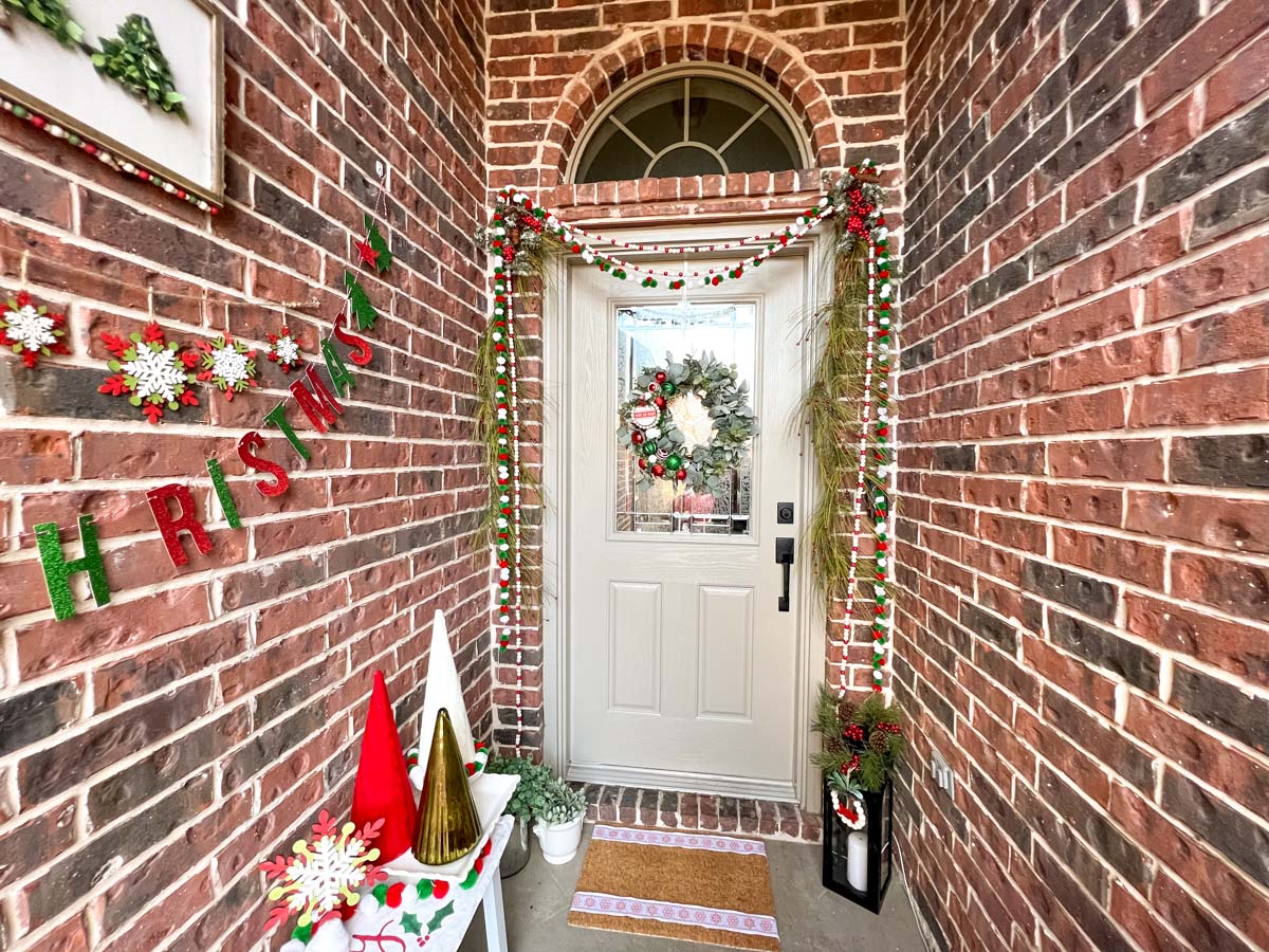 A tan front door with a holiday wreath of greenery and ornaments, garlands draping the door and a doormat decorated with Christmas ribbons. 