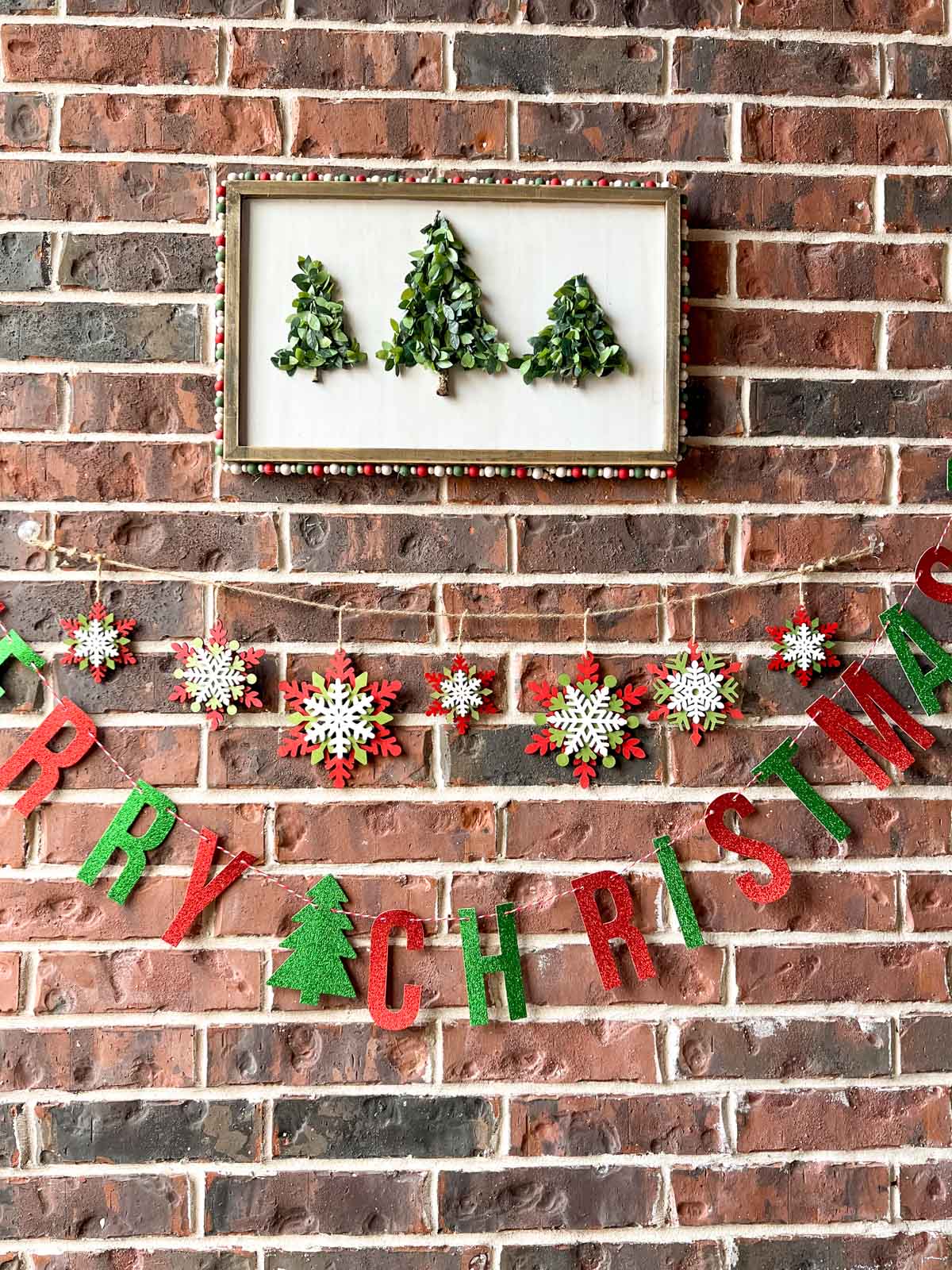 A wooden sign hanging on the wall that has 3 triangle shaped christmas tree made from greenery. The frame has christmas colored beads on the outside. Below the sign is a colored snowflake garland and a glittered "Merry Christmas" banner.