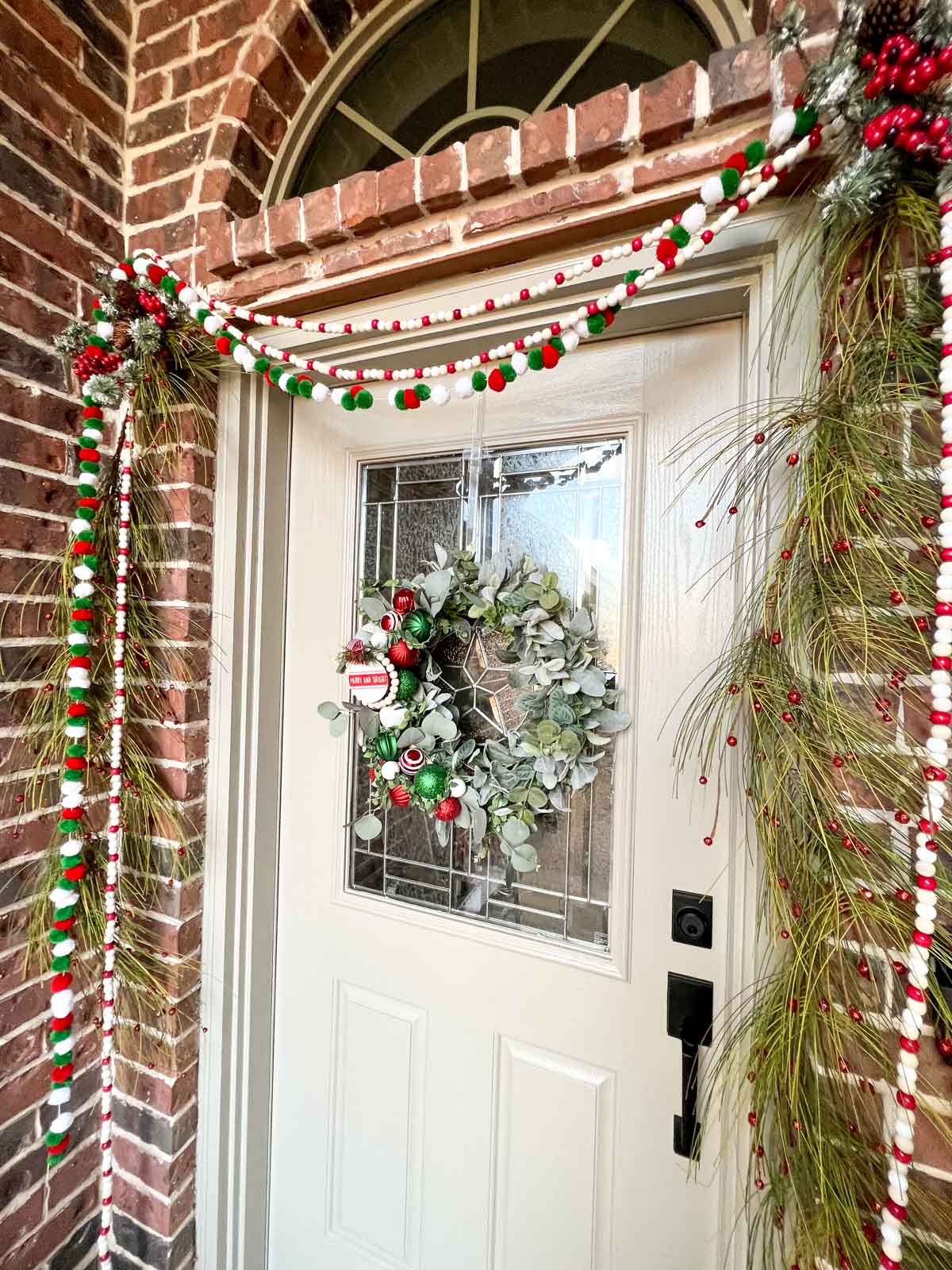a holiday wreath on a tan front door with pom pom and wooden garlands among evergreen garlands.