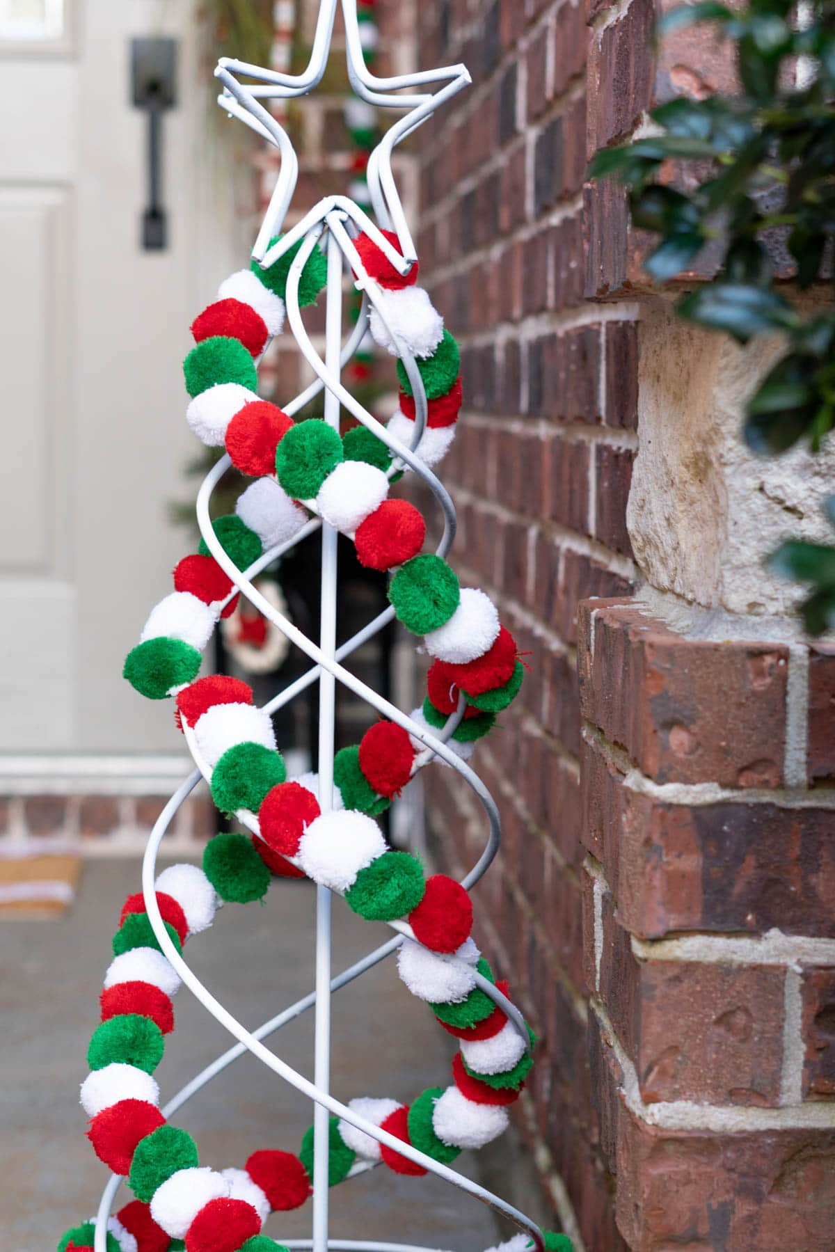 A white swirled wire Christmas tree is outfitted with a green, red, and white pompom garland on a front porch.