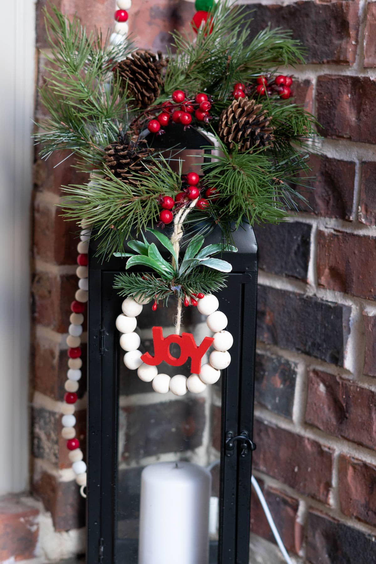 a basic black lantern with a holiday pine and berry floral candle ring placed atop with a beaded ornament draping off it.
