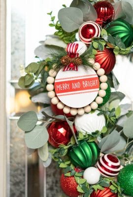A close up of red, green, and white Christmas ornaments of different sizes and textures on a lambs ear and eucalyptus wreath on a front door.