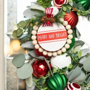 A close up of red, green, and white Christmas ornaments of different sizes and textures on a lambs ear and eucalyptus wreath on a front door.