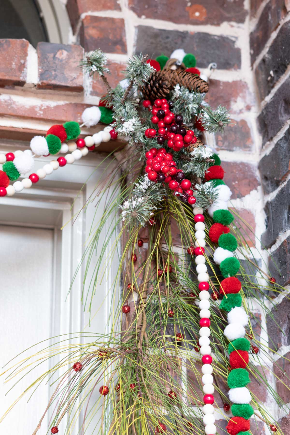 The corner of holiday garlands hung. A close up of a loose pine garland with red berries, a green, red and white pom pom string, and a red, white and natural wooden bead string, topped with frosted evergreens and red berries.