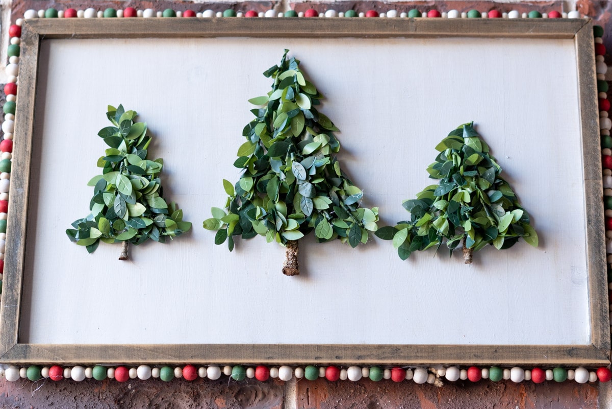 a wooden sign with 3 triangle christmas trees made from greenery garland. The wood frame has small colored beads around the edge.