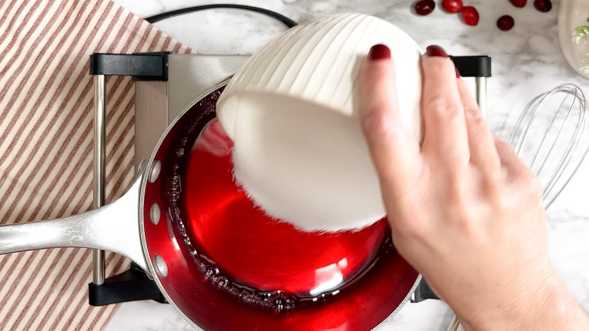 A small white bowl full of granulated sugar is being poured into a saucepan that has cranberry juice in it.