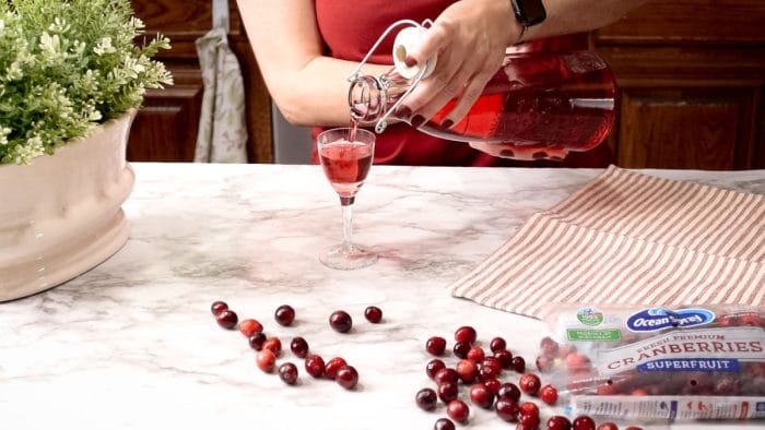Pouring cranberry cordial from a glass carafe into a stemmed vintage shot glass.