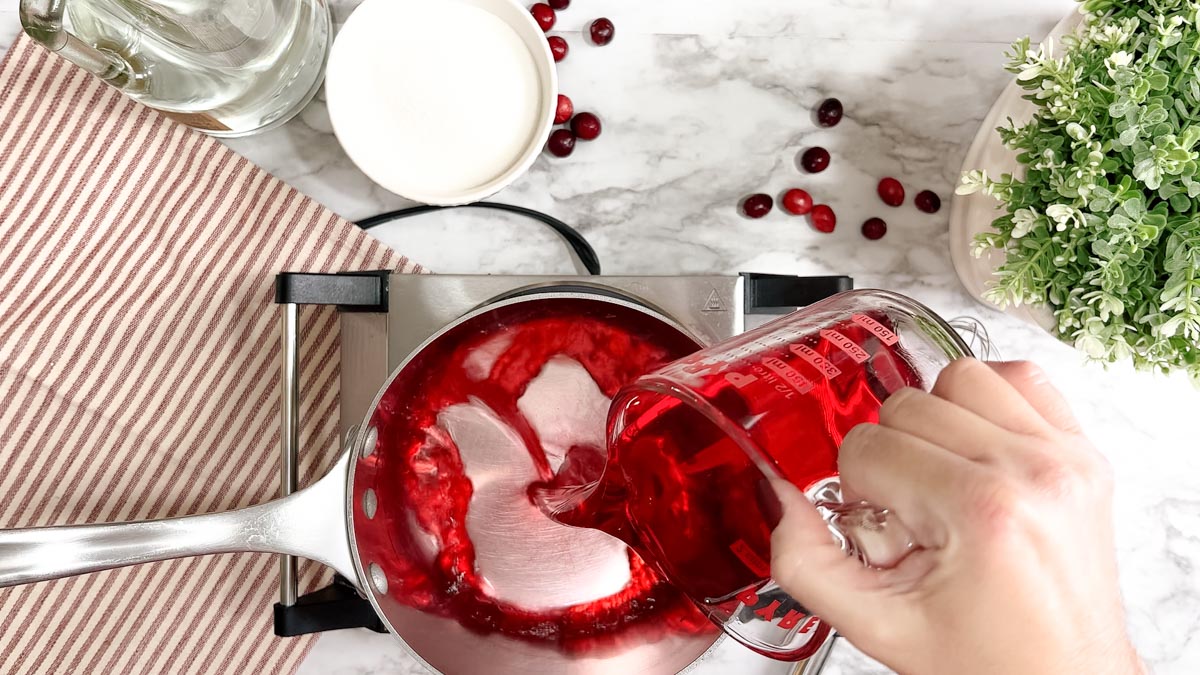 A glass measuring cup of cranberry juice cocktail being poured into a stainless steel saucepan on a burner.