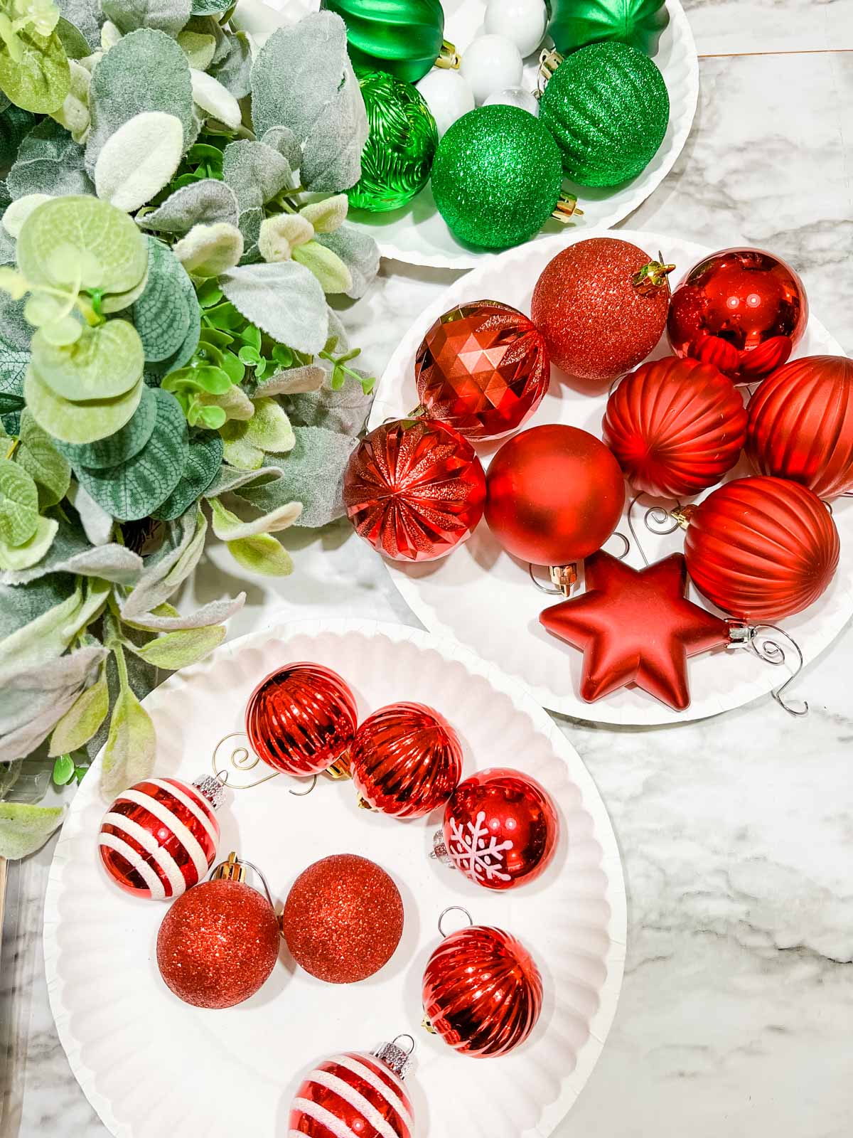 A variety of Christmas ornaments on plates next to a wreath in shades of red and green and in varying sizes.