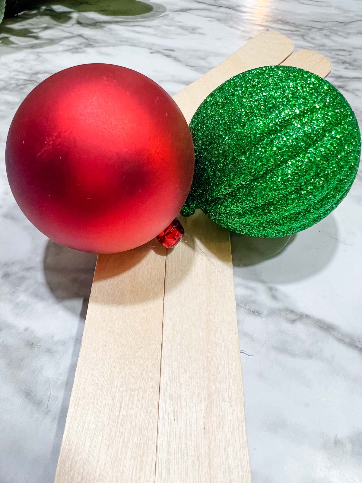 A solid red ornament and a green glitter ornament being hot glued to 2 large wooden popsicle sticks.