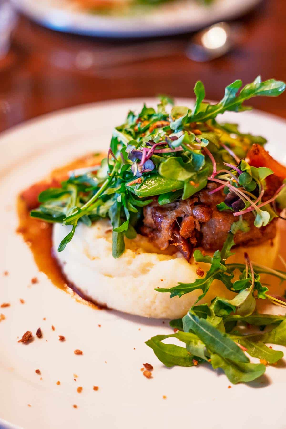 a white plate with sweet and sour piglet cheek with peas, apricots, fava beans, mustard pickles, fried garlic, and watercress on mashed potatoes.