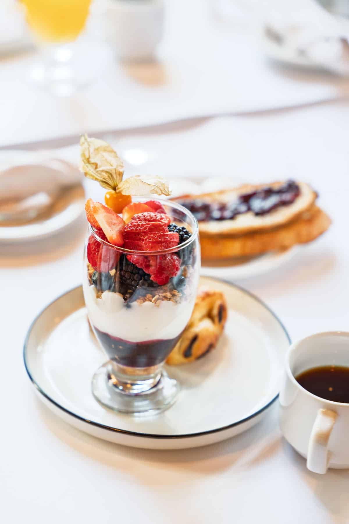 a white plate with a parfait glass filled with berry compote, vanilla yogurt, granola, and strawberries, blueberries and blackberries. Next to a cup of coffee, a pastry and a piece of jellied toast.