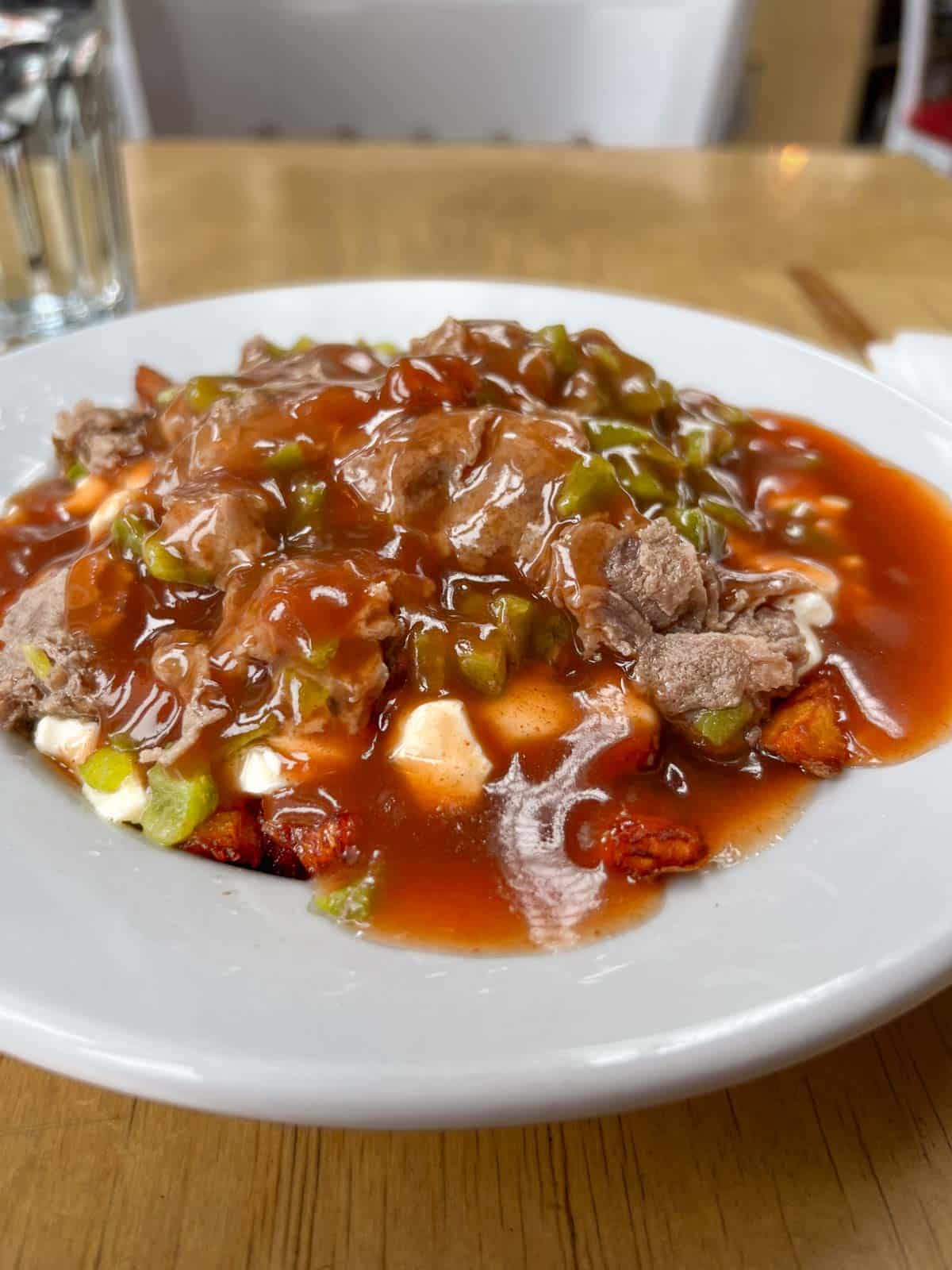 a white bowl filled with French fries, topped with green peppers, Philly steak, cheese curds and brown gravy.