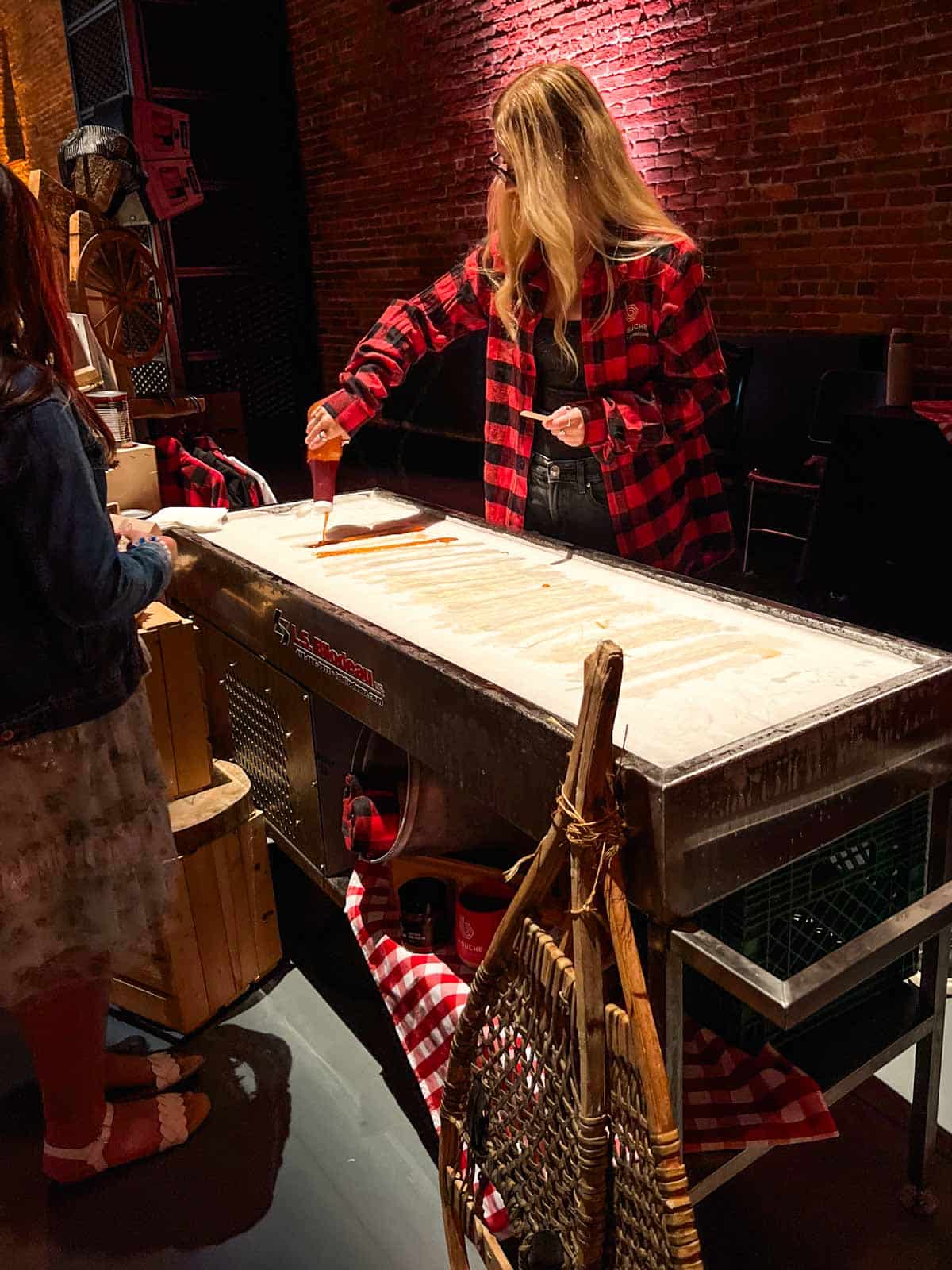 a girl in a plaid shirt pouring maple syrup from a bottle onto a bed of snow.