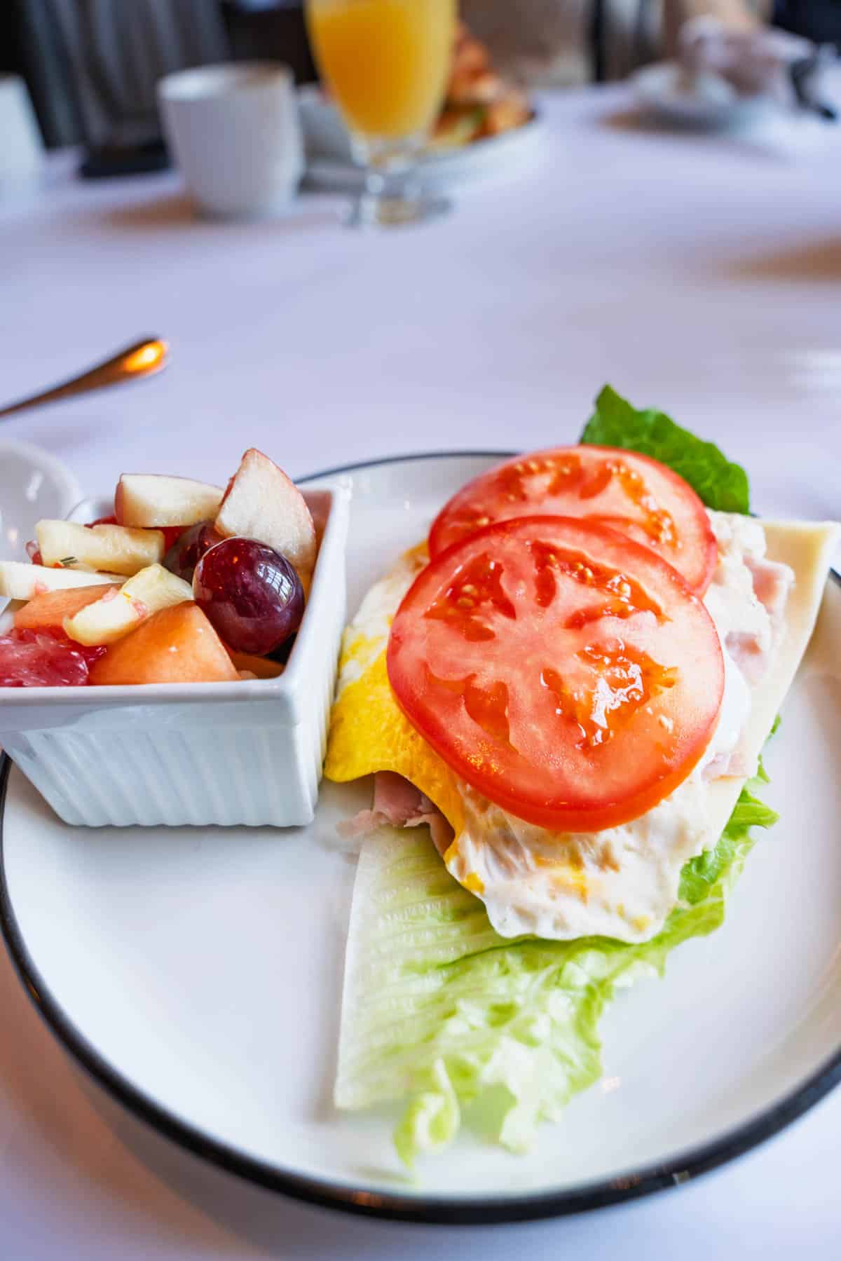 a white plate with layers of lettuce, ham, fried egg, and tomatoes on the right side, and a square bowl of mixed fruit on the left.