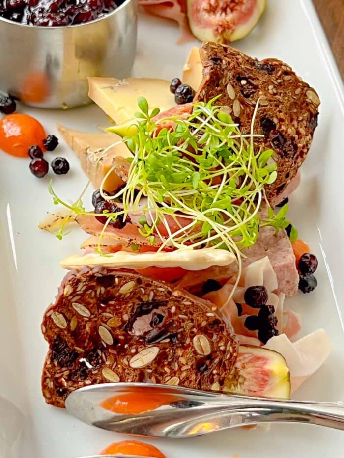 a white platter filled with charcuterie items: seeded toasted bread, meats, pates, assorted cheeses, micro greens, figs, and a silver container of blueberry chutney.