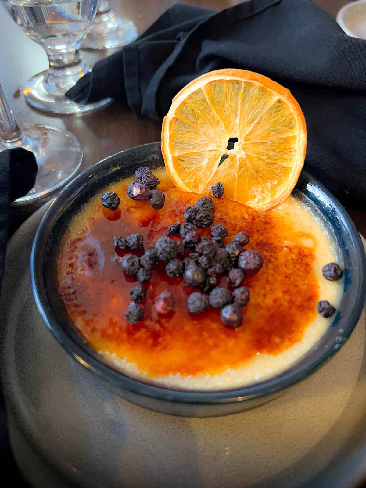 a short black bowl containing creme brûlée with a caramelized top, along with small blueberries and a dried orange slice.