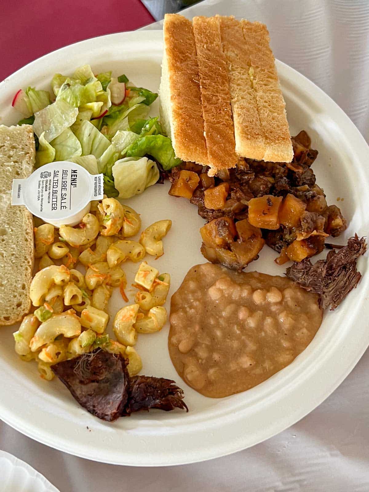 a white paper plate containing bread, a packet of butter, a salad, a mixture of moose meat and potatoes, macaroni salad, beaver meat, beans and, goose meat.